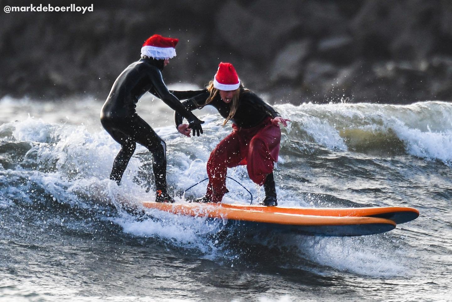 Two surfers wearing Santa hats ride a wave together on an orange surfboard.