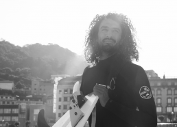 Surfer holding a board, standing outdoors with a serene expression. hoping for better surfboard fins
