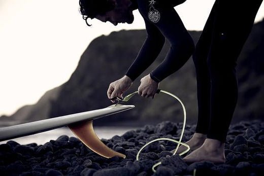 Surfer securing a leash to a surfboard on a rocky shoreline at dusk.