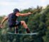 Man wakeboarding mid-air, wearing vibrant boardshorts and helmet, against a lush green backdrop.
